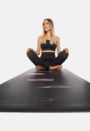 Zwarte yogamat met een gladde textuur, met paarse gradientpatronen. Model zit met gekruiste benen, gekleed in een cropped-top en leggings.