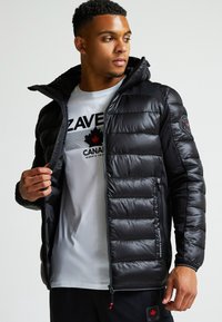 Čierna perová bunda s kapucňou, ktorá má prešitý dizajn, lesklý povrch a predný zips. Nosená cez svetlo šedé tričko s grafikou.