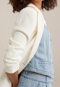 Biały blezer narzucony na niebieski dżinsowy kombinezon z guzikami. Blezer ma gładką fakturę; kombinezon charakteryzuje się luźnym krojem i kieszeniami.