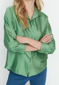 Grön satin knäppskjorta med krage, långa ärmar som är uppvikta vid manschetterna, och en något böjd nederkant. Lätt passform, slät textur.