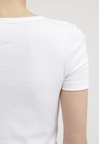 Vit kortärmad topp i ribbad bomull, med rund halsringning och en slät textur med minimala sömmar på baksidan.