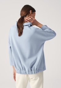 Felpa oversize azzurra chiara con un colletto alto, orlo arricciato e maniche lunghe, indossata sopra pantaloni bianchi con texture. Tessuto morbido.
