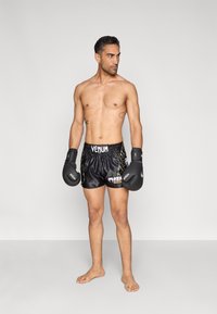 Μαύρο σατέν σορτς πυγμαχίας με κίτρινα στοιχεία, με το λογότυπο "VENUM" στη μέση; μαύρα γάντια πυγμαχίας; γυμνά πόδια; ουδέτερο γκρι φόντο.