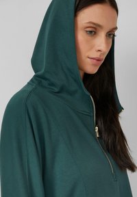 Felpa con zip e cappuccio in tessuto verde profondo, caratterizzata da un design leggero e da un dettaglio della zip visibile sulla parte anteriore.