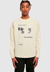 Langärmliger cremefarbener Sweatshirt mit bedruckten Grafiken: "HACKNEY E8", Anführungszeichen, "STAY SURREAL" und "apoh London" in Grau. Lockere Passform.