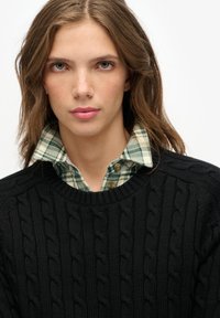 Zwarte gebreide kabeltrui met een ronde hals, gedragen over een groen-wit geruit overhemd met kraag, met knoopdetails.