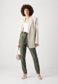 Beige oversized Leinenblazer, weißes Tanktop, olivgrüne Hochbundhose mit Schleife und helle Sandalen. Minimalistische Tasche in der Hand.