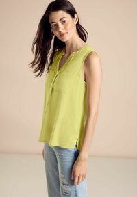 Top senza maniche di un vivace verde lime, realizzato in materiale leggero, con colletto e due tasche frontali. Indossato con jeans strappati.