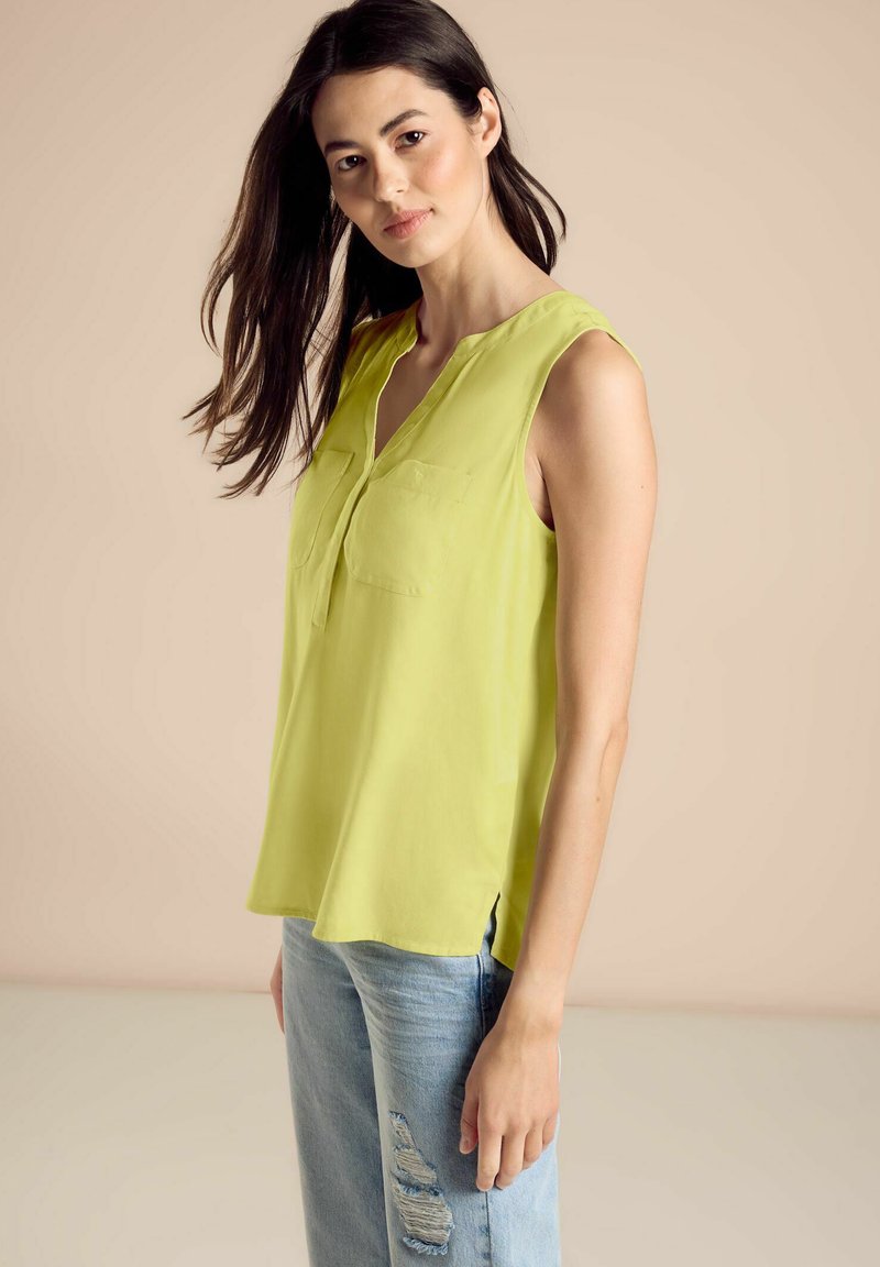 Top senza maniche di un vivace verde lime, realizzato in materiale leggero, con colletto e due tasche frontali. Indossato con jeans strappati.