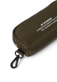 Olivgrön dragkedjefodral gjort av texturerad tyg, med en svart klämma och vitt präglat bokstäver på ena sidan. Rektangulär form.