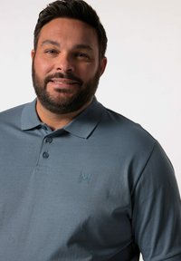 Šedé polo tričko vyrobené z měkkého materiálu, s tříknoflíkovou légou a jemným logem na hrudi. Hladká textura bez vzorů.