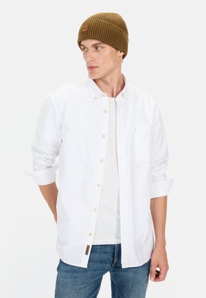 White button-up shirt layered over a white t-shirt, paired with a brown ribbed knit beanie. Blue denim jeans complete the look.