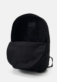 Sac à dos noir de forme arrondie, fabriqué en tissu durable. Il comporte une fermeture à rabat et une poche intérieure avec un bord froncé.
