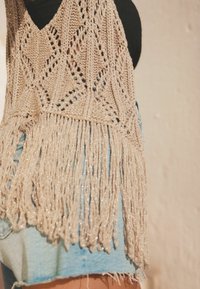 Pletený béžový top bez rukávů s diamatovým vzorem a třásněmi na lemu. Spárován se světle modrými džíny s ošuntělými okraji.