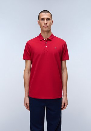 Junger Mann steht, trägt ein rotes Poloshirt mit kurzen Ärmeln und dunkelblaue Hose vor einem schlichten hellen Hintergrund.