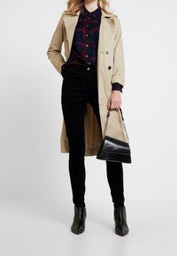 Beige trenchcoat, marineblauw geruit overhemd met knoppen, zwarte skinny broek, zwarte hakken enkellaarzen, en een kleine zwarte handtas met kettinghengsel.