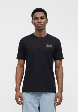 Mand iført en sort t-shirt med korte ærmer med et EA7 Emporio Armani-logo på venstre bryst og lyseblå jeans.