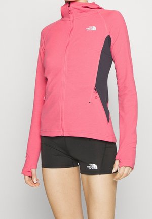 Femme portant une veste rose zippée et un short noir avec les logos The North Face, debout devant un fond clair uni.