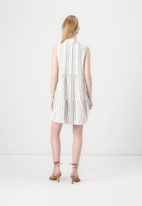Femme aux longs cheveux blonds porte une robe blanche sans manches à rayures verticales et des talons marron à brides, debout contre un mur blanc uni.