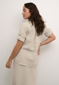 Lichtbeige linnen shirt met opgerolde mouwen, subtiele achternaaddetail en een relaxte pasvorm, gedragen over een bijpassende rok, staande tegen een effen achtergrond.