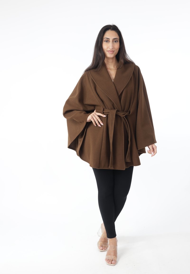 Abrigo envolvente marrón de gran tamaño con un cuello ancho, cinturón de lazo y tela texturizada, combinado con leggings negros y sandalias transparentes de tacón.