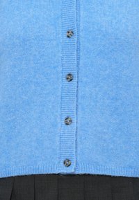 Lichtblauwe cardigan van zacht materiaal, met een ribbelstructuur en zes donkere knopen in het midden. De onderrand is ribbelig.