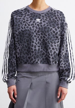 Person wearing a gray Adidas leopard-print sweatshirt with white stripes on sleeves and a gray pleated skirt.