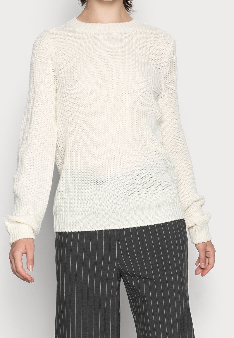Femme portant un pull en maille blanc et un pantalon à fines rayures gris foncé, debout devant un fond uni.