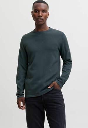 Homme portant un pull texturé à manches longues vert foncé et un pantalon noir, debout avec une main dans la poche sur un fond uni.