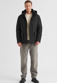 Schwarzer isolierter Parka mit Kapuze, Reißverschluss vorne und Druckknopfverschlüssen. Zwei Fronttaschen; getragen über einem cremefarbenen Pullover und grauen Jeans.