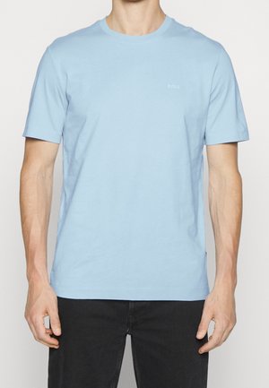 Mann trägt ein schlichtes hellblaues kurzärmeliges T-Shirt und schwarze Hosen, steht vor einem schlichten weißen Hintergrund.