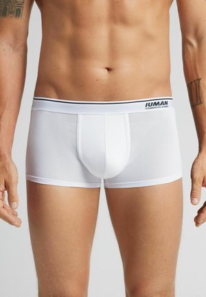 Männlicher Oberkörper in weißen Boxerunterhosen mit einem markierten Bund, sichtbare tätowierte Arme, steht vor einem neutralen Hintergrund.