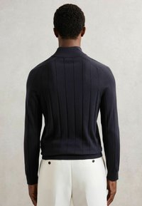 Granatowy sweter roboczy z pionowym żebrowaniem, wysokim kołnierzem i dopasowanymi mankietami, w połączeniu z białymi spodniami z dwoma widocznymi guzikami z tyłu.