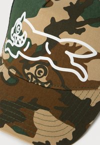 Camouflage baseball cap with a white outlined cartoon cat design featuring sharp teeth and spiral eyes on the front panel.
