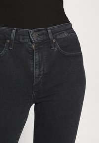 Jeans de mezclilla negros de talle alto, combinados con una blusa negra metida por dentro, que cuenta con bolsillo delanteros, pasadores para cinturón, botón y cierre de cremallera.