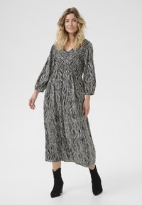 Pitkä musta-valkoinen kuviollinen mekko, jossa on puffihihat, V-kaula-aukko ja kiristetty vyötärö, yhdistettynä mustiin nilkkureihin.