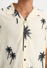 Camisa de manga corta, de color beige claro, con cuello, que presenta un patrón de palmeras negras y tres botones en la parte delantera. Textura de tejido suave.