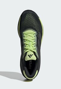 Zapato atlético negro con parte superior de malla, detalles en verde lima y suela redondeada. Presenta cordones prominentes y área de punta reforzada.