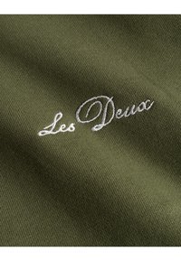 Tessuto verde oliva con logo ricamato "Les Deux" in testo corsivo bianco, che presenta una texture liscia e una leggera lucentezza.