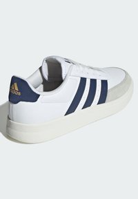 Bijela tenisica s tamnoplavim tri pruga, nubuk kapicom, teksturiranom gumiranom potplatom i zlatnim logom na peti, s vezivanjem na vezice.