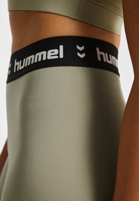 Legginsy w oliwkowym kolorze o gładkiej fakturze i czarnym elastycznym pasku w talii z białym logo "hummel" i wzorem w chevron.
