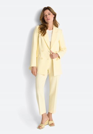 Žena s dugom kosom stoji noseći blijedo žuti dvoredni sako, odgovarajuće skraćene hlače, bijelu majicu i cipele s otvorenim prstima.