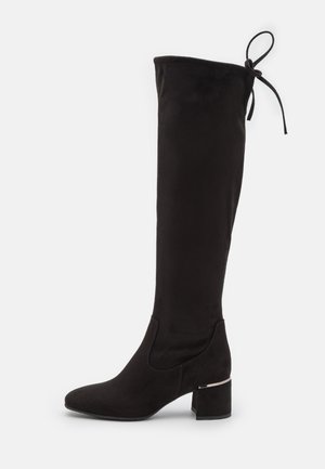 Botte noire montant jusqu'au genou en daim doux, avec un bout arrondi, un talon bloc et un détail noué en haut. Ornée de détails métalliques.