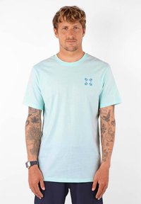 T-shirt bleu clair à col rond et à manches courtes. Présente un petit motif graphique bleu sur la partie gauche de la poitrine. Texture de tissu douce.