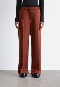 Rust-colored wide-leg trousers with a smooth texture, elastic waistband, front pleats, and split hems, paired with black shoes.