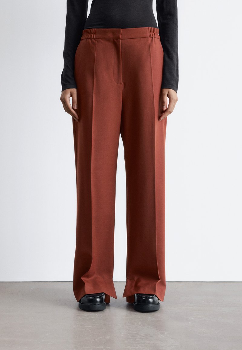 Rust-colored wide-leg trousers with a smooth texture, elastic waistband, front pleats, and split hems, paired with black shoes.