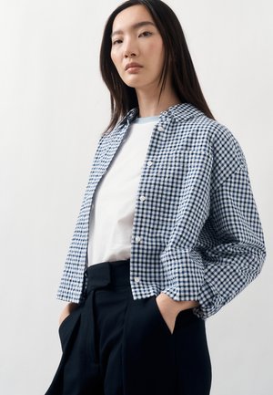 Młoda kobieta ubrana w niebiesko-białą koszulę w kratę na białym t-shircie, czarne spodnie, ręce w kieszeniach, na jednolitym tle.