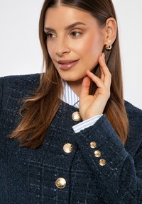 Námořnický texturovaný blejzr se zlatými knoflíky, se bílým a modrým pruhovaným límcem a manžetami. Hladké vlasy a jemný make-up, ruka na obličeji.