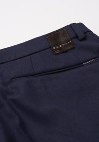 Navy blue trousers made of cotton blend fabric, featuring a back pocket, stitching details, and a rectangular brown label with branding.