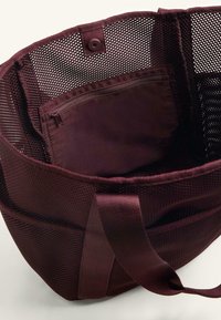 Bordowa siatkowa torba tote z materiałowymi akcentami. Wewnątrz znajduje się zasuwana kieszeń, wzmocnione uchwyty oraz zapięcie na zatrzask.
